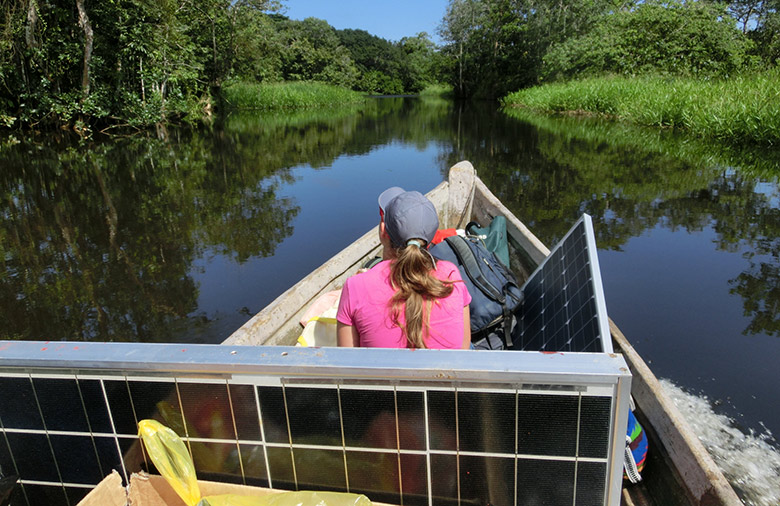 On voit de dos une femme sur un bateau qui navigue sur une petite rivière entourée d'une forêt dense. Deux panneaux solaires sont posés sur le bateau.
